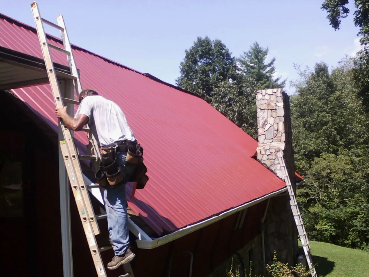 Roofer on a ladder beside a red metal roof and stone chimney — Soffit & Fascia Repair by Roofing Brothers in Gallatin Gateway